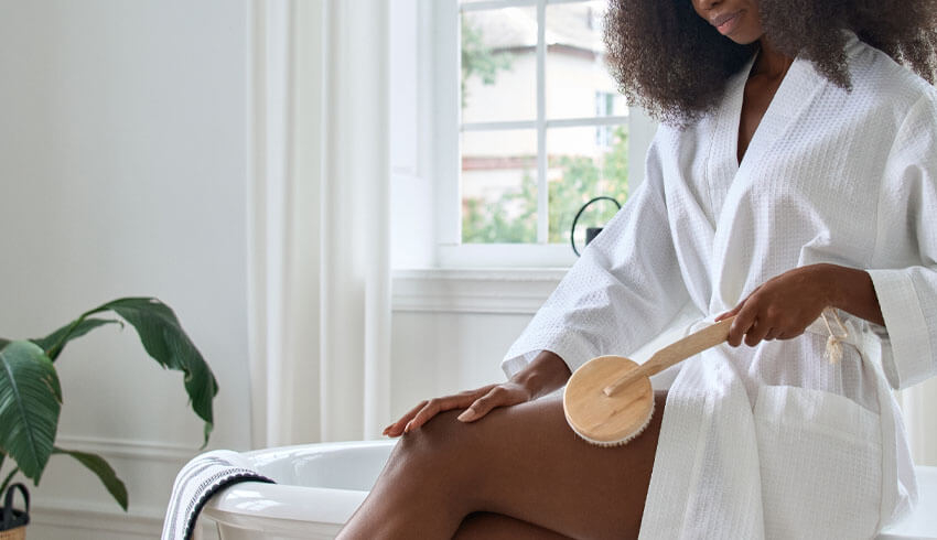 A woman wearing a bathrobe using a dry brush on her thigh